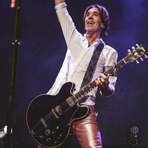 A musician wearing a white shirt and shiny pink pants plays an electric guitar on stage, smiling and raising one hand triumphantly into the air under purple stage lights, reminiscent of the iconic energy of Roxette band members.