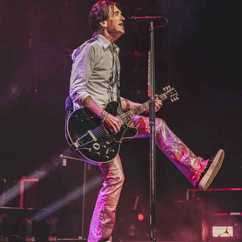 A musician, reminiscent of Roxette band members, plays an electric guitar and sings into a microphone on stage, lifting one leg energetically. Stage lights and a smoky atmosphere create a lively concert scene.