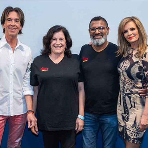 Four adults, resembling Roxette band members, stand side by side, smiling at the camera. Two are in casual tops and jeans, while the others are dressed more formally. They are posed against a blue and white wall.