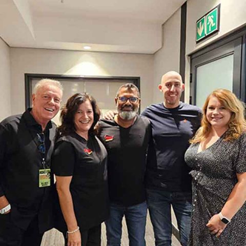 Five smiling people, resembling Roxette band members, stand together indoors in a hallway for a group photo. They are casually dressed and close together, with glass doors and an emergency exit sign in the background.
