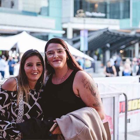 Two women, reminiscent of Roxette band members, stand closely together and smile at the camera outside at an event, with a fenced-off area, people, and buildings visible in the background.