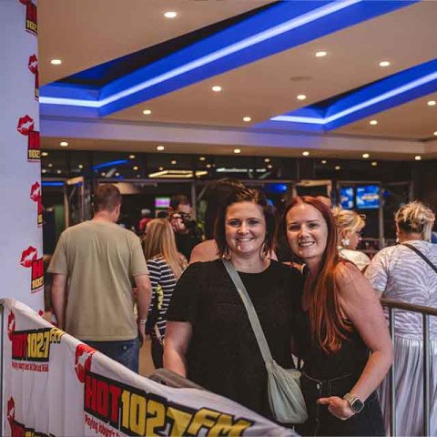 Two women smile at the camera while standing in front of a HOT 102.7 FM barricade at an indoor event, where Roxette band members music plays and people gather under bright blue ceiling lights.