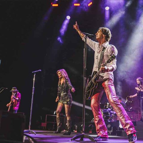 A band performs on stage under colorful lights; the lead singer in shiny pants, reminiscent of Roxette band members, plays guitar and raises one arm, while other band members sing and play instruments.