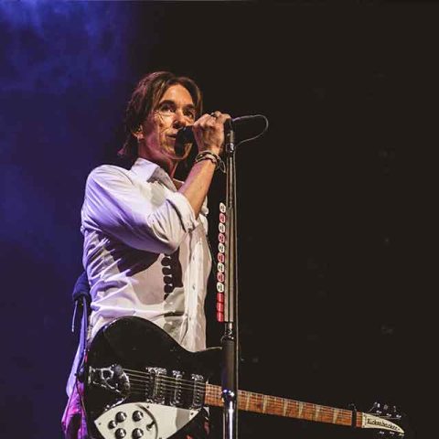 A musician in a white shirt sings into a microphone on stage, holding a black electric guitar, with purple stage lighting and a dark background, reminiscent of Roxette band members performing live.