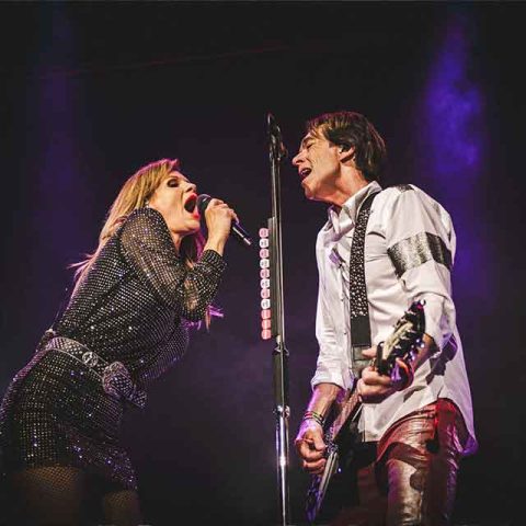 A woman sings passionately into a microphone while a man beside her, resembling Roxette band members, plays guitar and sings, both performing on stage under dramatic lighting.