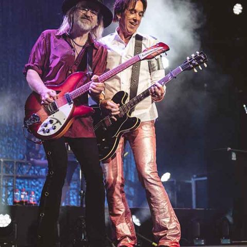 Two male Roxette band members perform on stage, each playing electric guitars. One wears a hat, sunglasses, and a maroon shirt; the other sports a white shirt and shiny pink pants. Stage lights and smoke create a dynamic atmosphere.