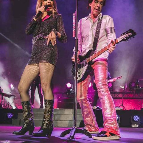 A woman, one of the Roxette band members, sings into a microphone while a man in shiny pants plays electric guitar on stage, both performing under colorful lights with their band in the background.