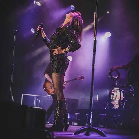 A female singer in a glittery outfit performs on stage, channeling the energy of Roxette band members. Purple stage lights shine around her as she holds a microphone, with a guitarist and drummer playing in the background.