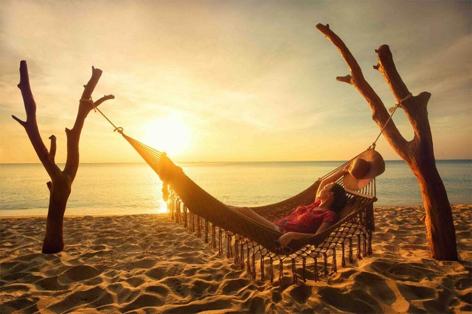 A person relaxes in a hammock tied between tree trunks on a sandy beach, watching the sunset over calm ocean waters, with a hat hanging on one trunk.