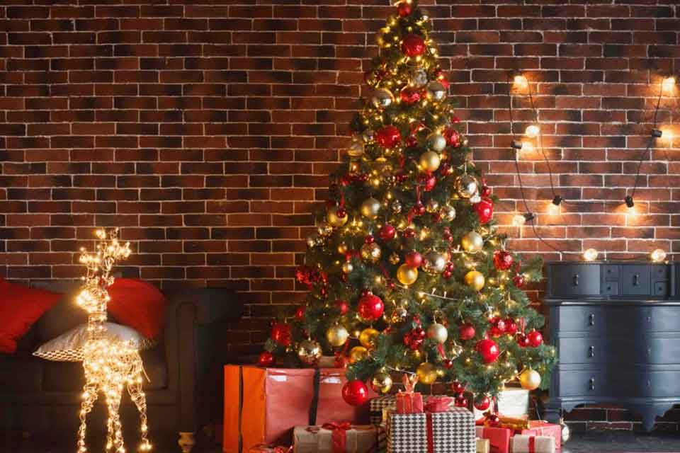 A decorated Christmas tree with red and gold ornaments stands in front of a brick wall, surrounded by wrapped gifts. A glowing reindeer decoration is on the left, and fairy lights hang on the right.