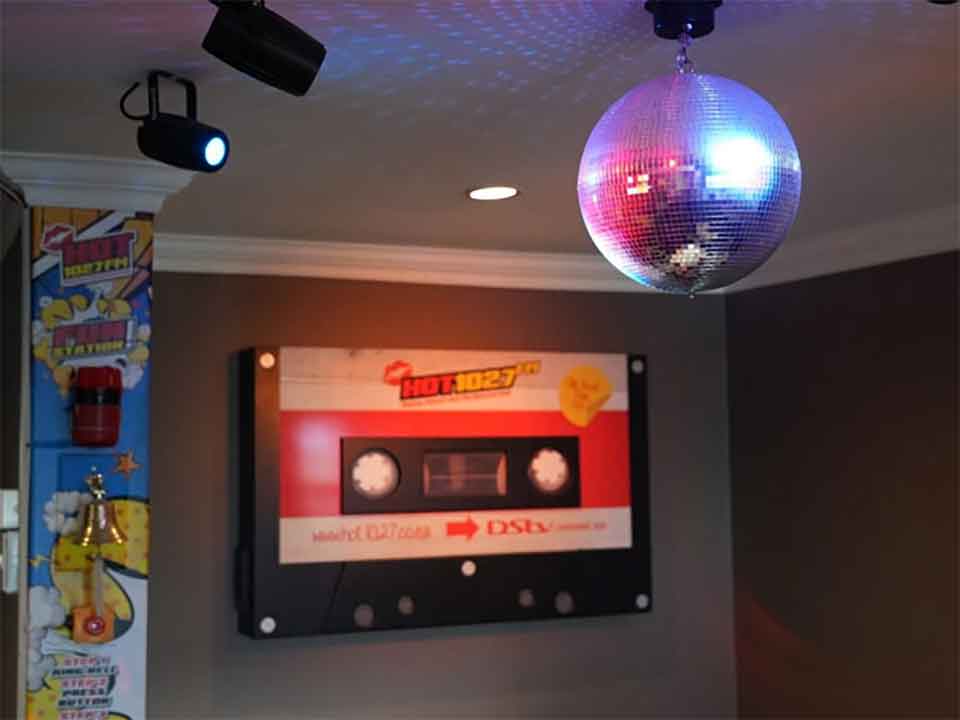 A shiny disco ball hangs from the ceiling, reflecting lights. On the wall, there is a large decorative cassette tape and colorful comic-style art on the corner pillar.