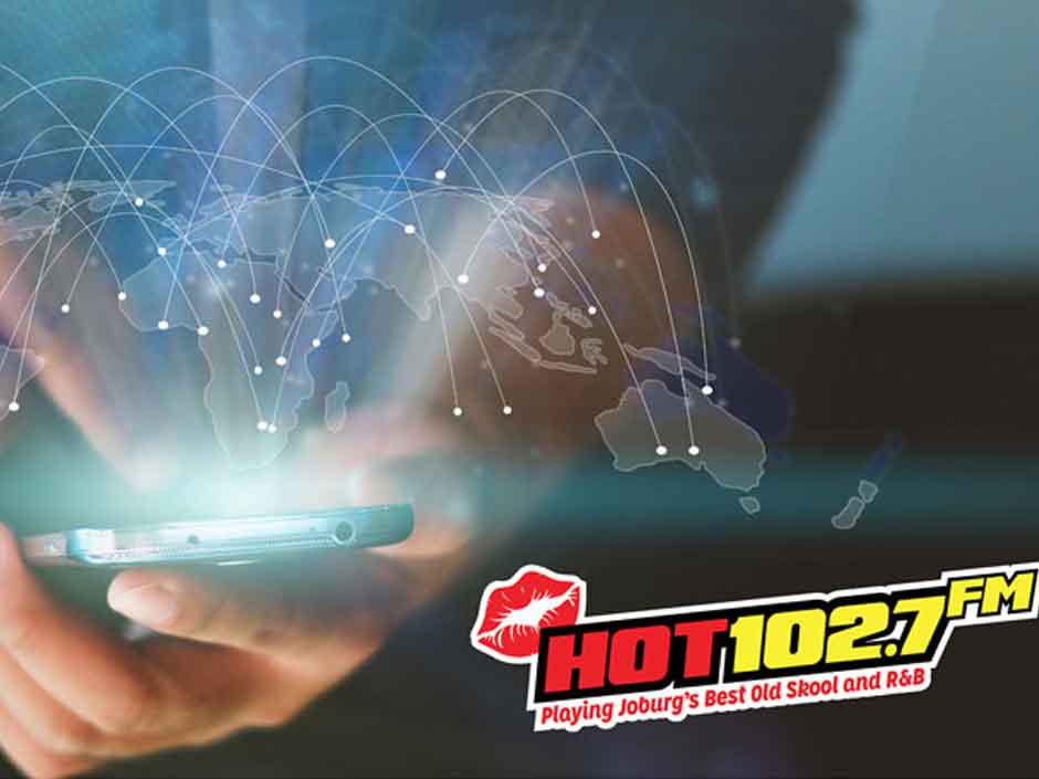 A person uses a smartphone with a digital world map and network lines overlay. The HOT 102.7 FM radio logo appears in the bottom right corner with the slogan, Playing Joburgs Best Old Skool and R&B.