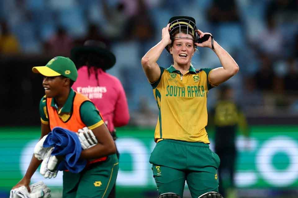 Two South African women cricketers celebrate on the field; one is holding her helmet with a joyful expression, while the other carries equipment. Both wear green and yellow uniforms.