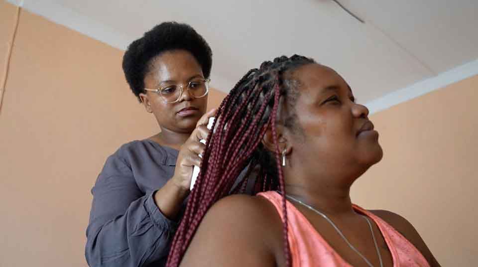 A woman with glasses styles another womans long red braids in a room with peach-colored walls. The seated woman wears a pink top and looks relaxed while her hair is being done.