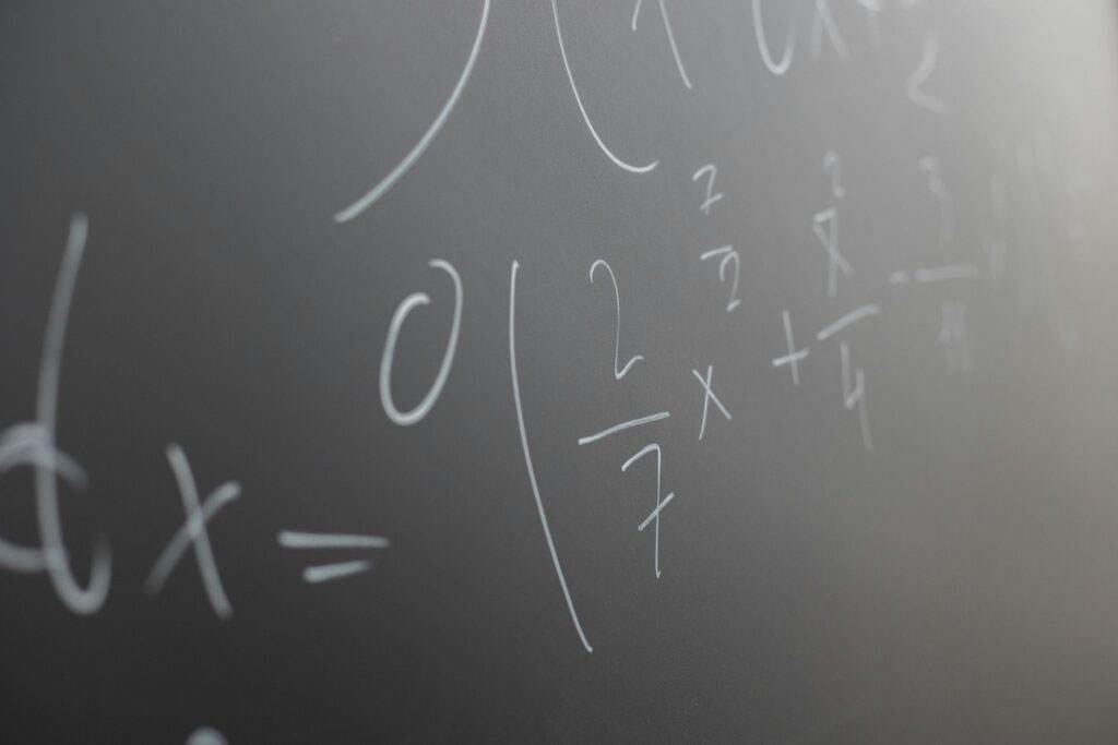 Mathematical equations written in white chalk on a blackboard, featuring fractions, variables, and parentheses. The focus is on the central part of the equations, with the rest fading out of focus.