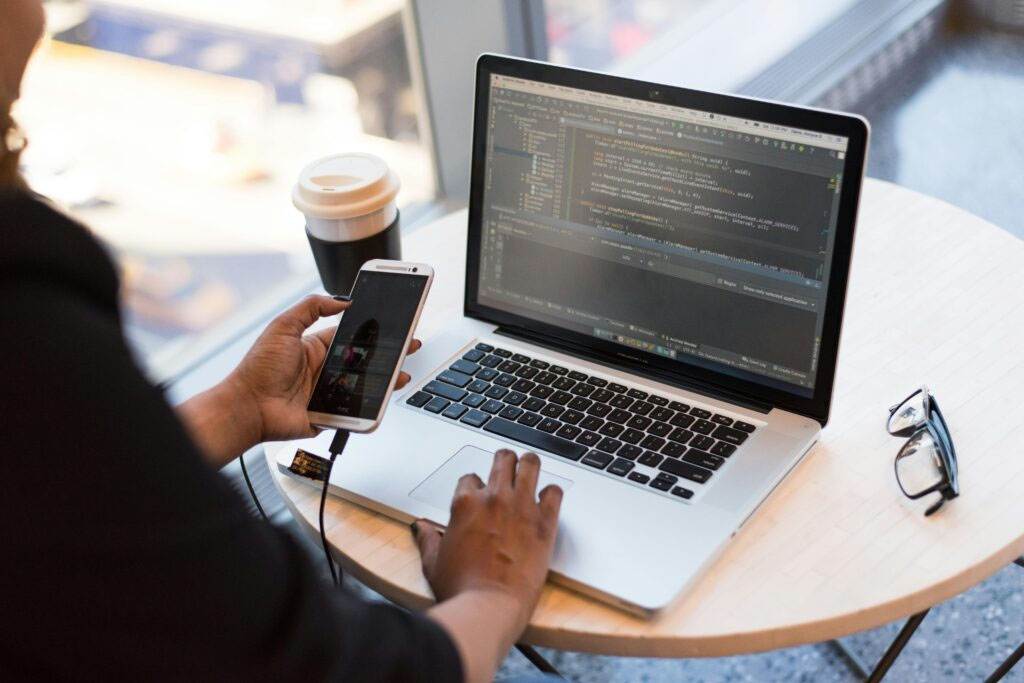 A person works on a laptop displaying code, while holding a smartphone connected to the laptop. A takeaway coffee cup sits on the small round table beside the laptop.
