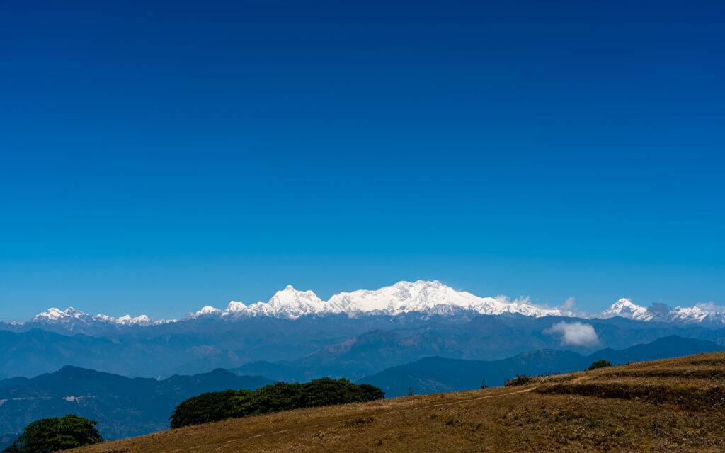 A clear blue sky above a distant range of snow-capped mountains, with green hills and brown grassy land in the foreground.