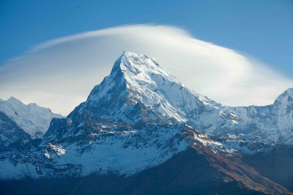 A snow-covered mountain peak rises sharply against a blue sky, with a wispy, white lenticular cloud hovering above it. The lower slopes have patches of exposed rock and sparse snow.