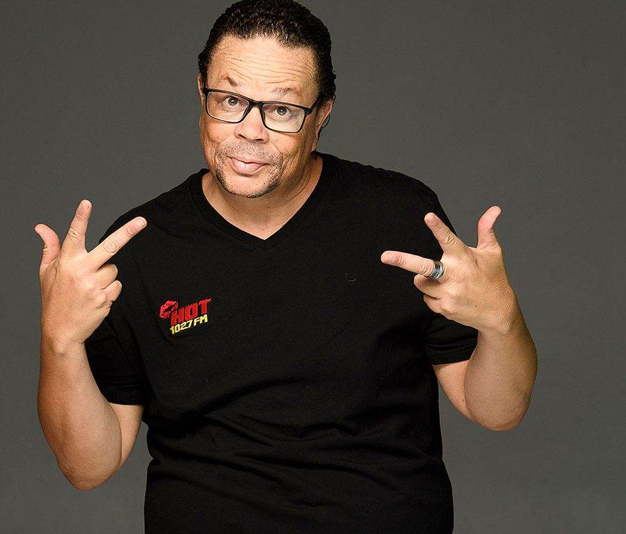 A man with glasses and short curly hair wearing a black Hot 102.7 FM t-shirt poses against a plain gray background, holding up both hands with his index and middle fingers extended in a peace sign gesture.
