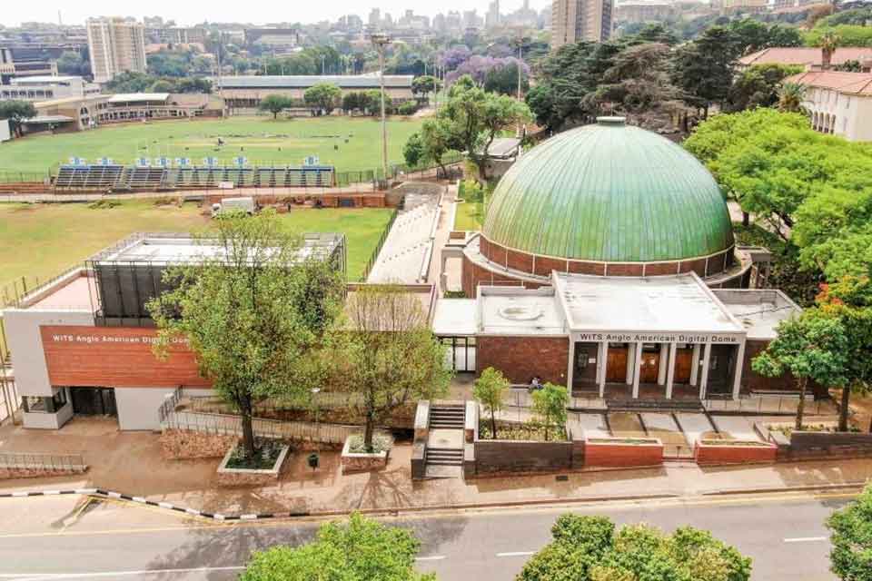 Aerial view of WITS Anglo American Digital Dome and surrounding buildings, with a green dome roof, trees, a sports field, and a cityscape in the background.