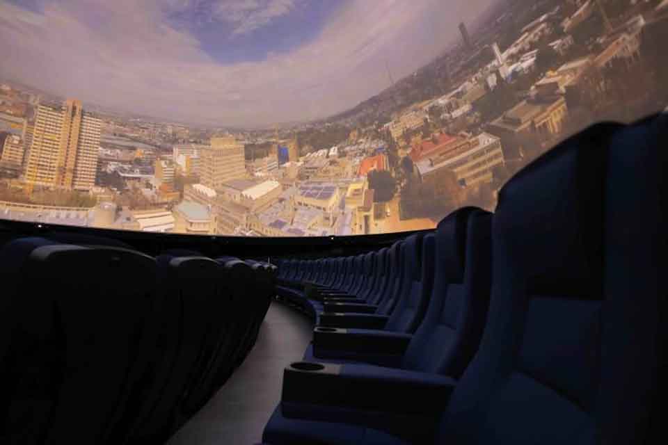 A row of empty, dark theater seats faces a large curved screen displaying an aerial view of a city under a cloudy sky, suggesting an immersive or planetarium-like experience.