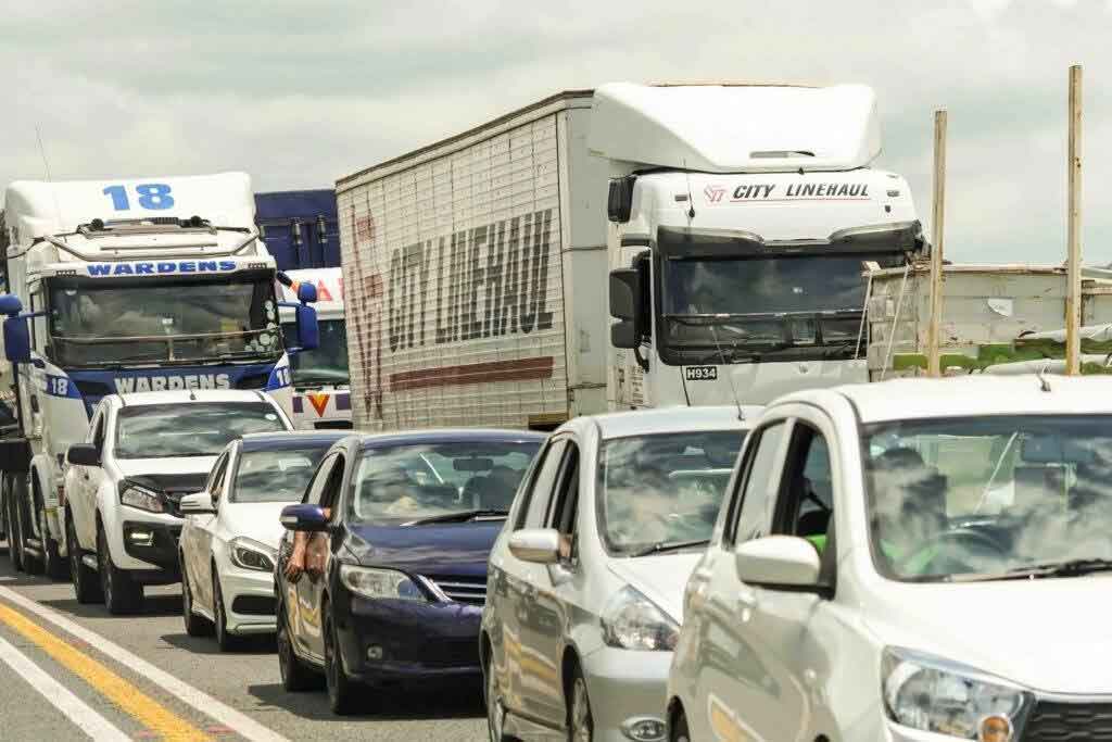A line of cars and trucks stuck in traffic on a road during the daytime, with some vehicles facing the camera and others moving in the opposite direction. The sky is cloudy.
