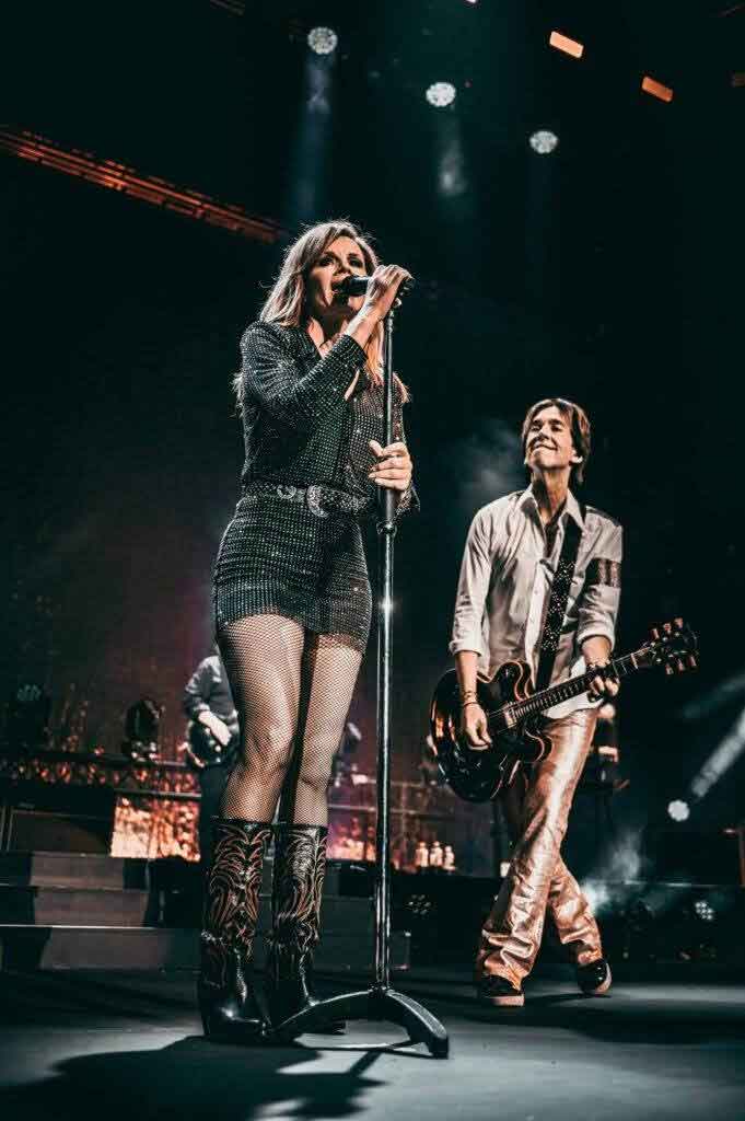 A woman sings into a microphone while a man next to her plays guitar on stage. Both are illuminated by stage lights, with band members and instruments in the background. The atmosphere suggests a live concert performance.