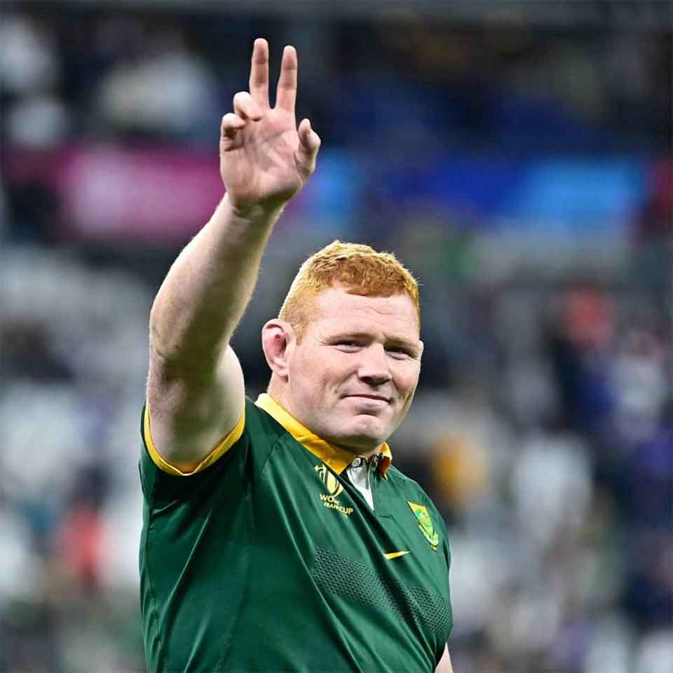 A rugby player in a green jersey with yellow trim raises his hand and gestures with two fingers. He has short red hair and is standing in a stadium with a blurred crowd in the background.