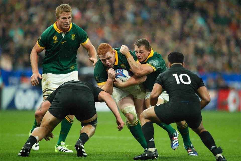 A rugby player in a green jersey runs with the ball while being tackled by two players in black jerseys, as another teammate in green supports him. The action takes place on a grass field during a match.