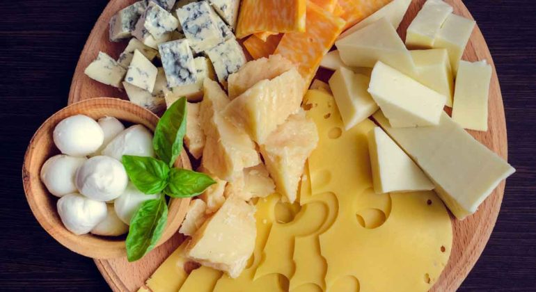 A round wooden board with assorted cheeses, including Swiss, mozzarella balls, blue cheese, cheddar, colby jack, parmesan wedges, and garnished with fresh basil leaves, on a dark surface.