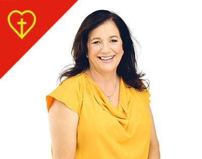 A woman with long dark hair wearing a yellow top smiles at the camera. In the top left corner, there is a red triangle with a yellow heart and cross symbol inside. The background is white.