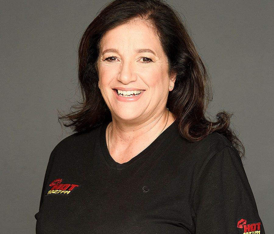 A woman with long dark hair smiles at the camera. She is wearing a black t-shirt with a red and yellow logo on the left side. The background is plain gray.