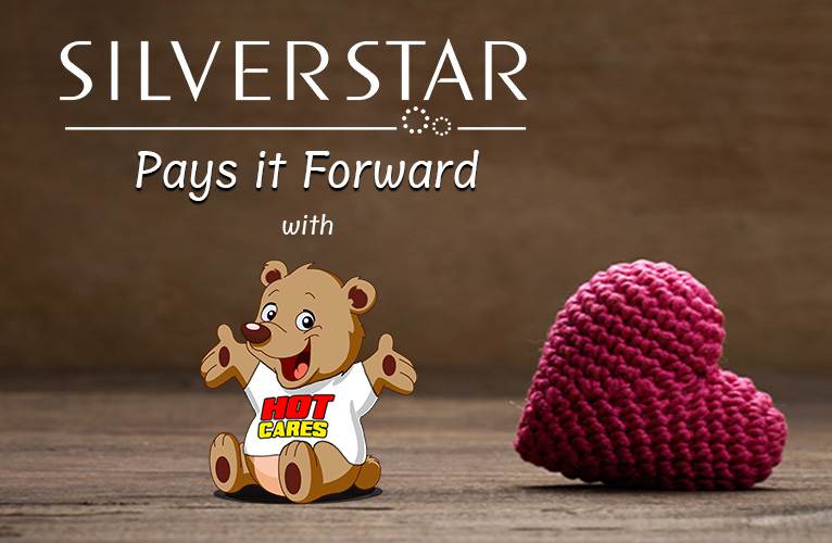 A brown teddy bear wearing a Hot Cares shirt sits next to a knitted red heart on a wooden surface. Above them, the text reads: Silverstar Pays it Forward with Hot Cares.