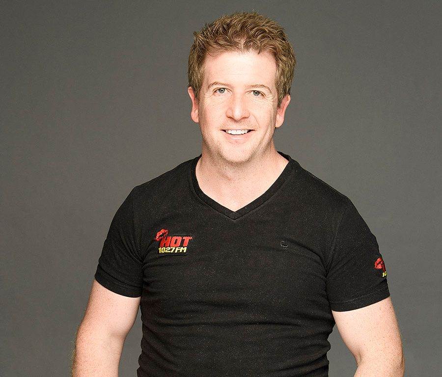 A man with short, light brown hair smiles at the camera. He is wearing a black t-shirt with HOT 102.7FM logos on the chest and sleeve, standing against a plain gray background.