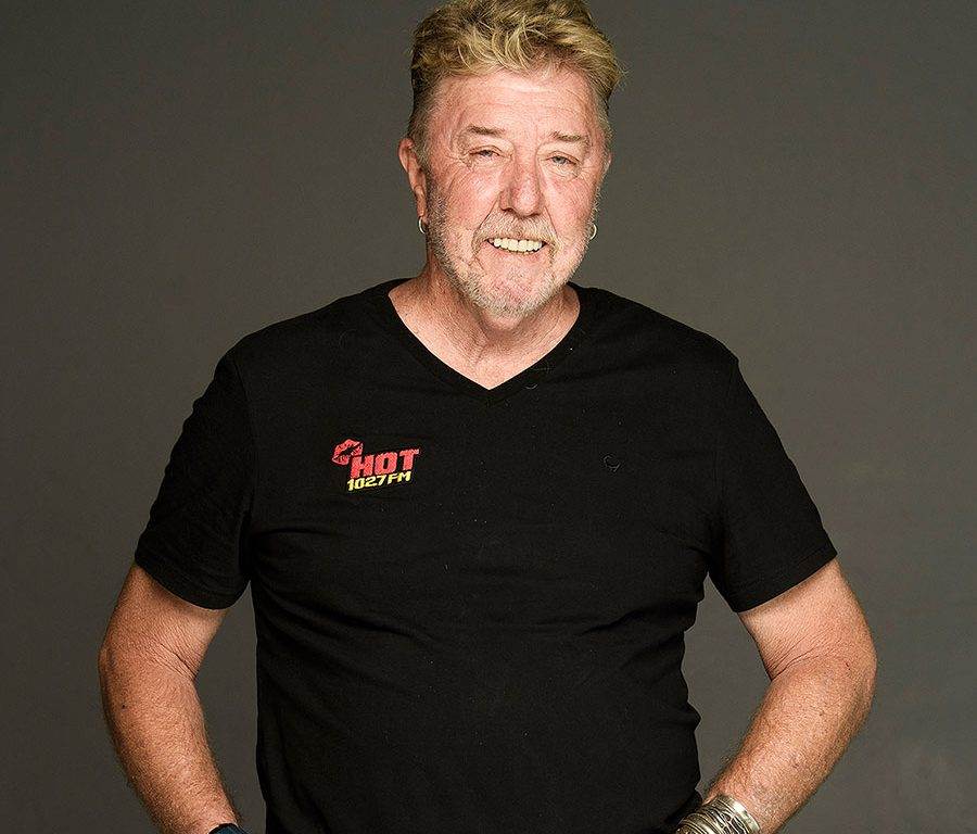 A smiling older man with short, light hair and a beard stands against a plain background. He wears a black t-shirt with a red HOT 102.7 FM logo and has his hands on his hips. He is also wearing earrings and bracelets.