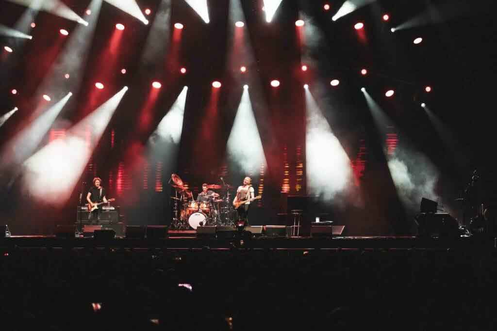 A rock band performs on stage under bright red and white lights, with smoke effects and musical instruments visible; a dark audience is in the foreground.