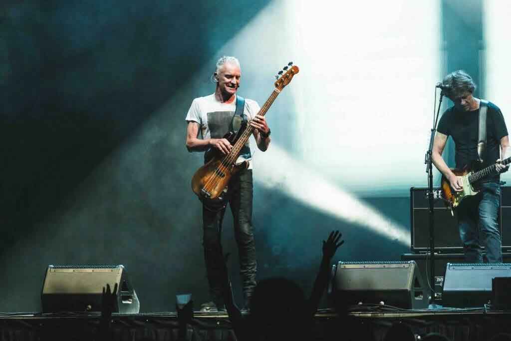 Two musicians perform on stage under dramatic lighting; one plays a bass guitar and sings, while the other plays an electric guitar. Fans’ silhouettes and raised hands are visible in the foreground.