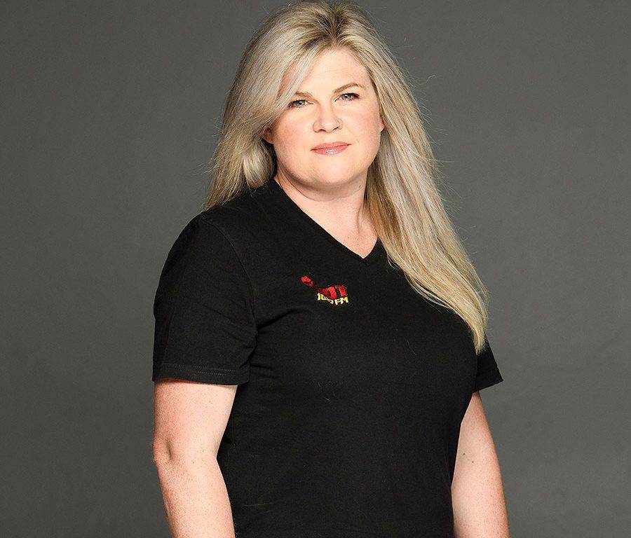 A woman with long blonde hair wearing a black V-neck T-shirt stands against a plain gray background, looking at the camera with a neutral expression.