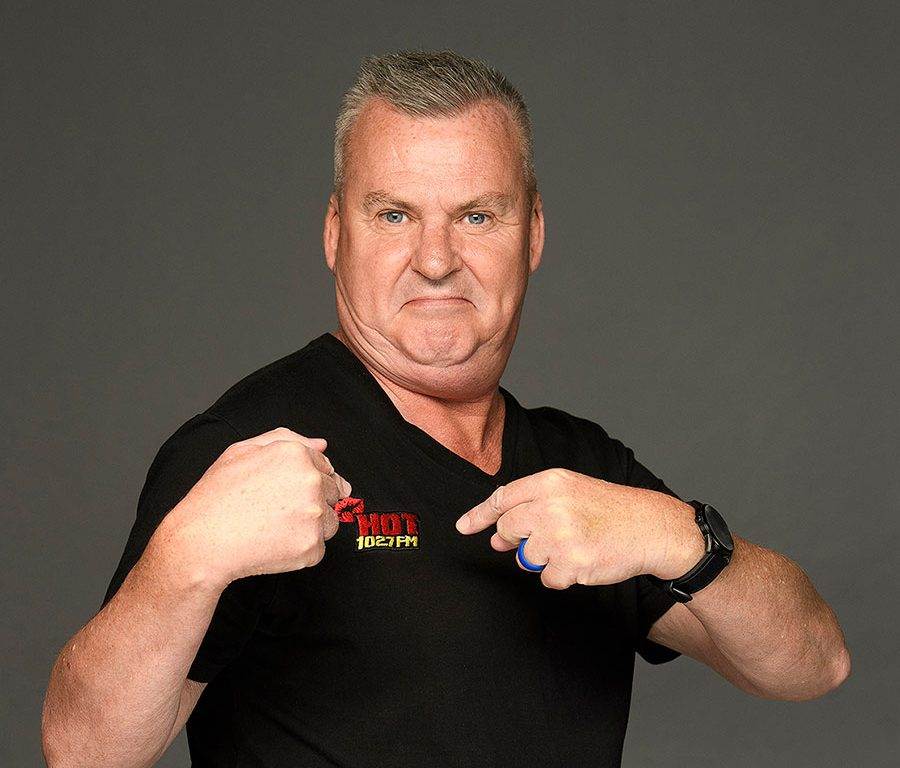 A middle-aged man with short gray hair wears a black t-shirt that says HOT 102 FM and points at the logo with both hands, standing against a plain gray background.