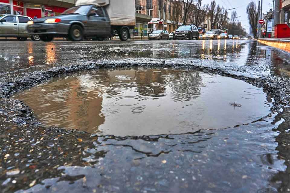 A large pothole filled with rainwater on a city street, with cars driving by and buildings in the background. Raindrops are creating ripples in the water.