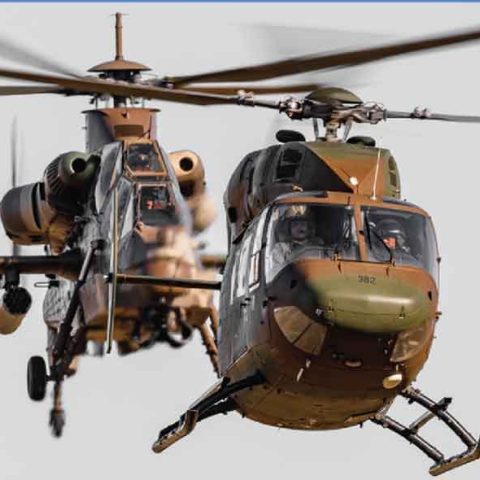 Two military helicopters in flight, with camouflage paint, fly closely together against a plain sky. The helicopter in the foreground is facing forward, while the one behind appears to be at an angle.