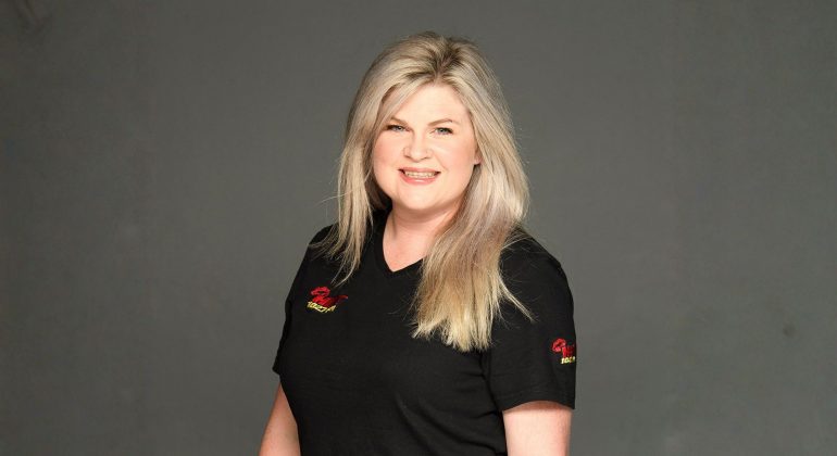 A woman with long blonde hair wearing a black t-shirt stands smiling in front of a plain gray background, celebrating her recent recognition at the New York Festivals Radio Awards.
