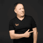 A middle-aged man with short hair wearing a black t-shirt stands against a dark background, smiling slightly and pointing to his left with his right hand.