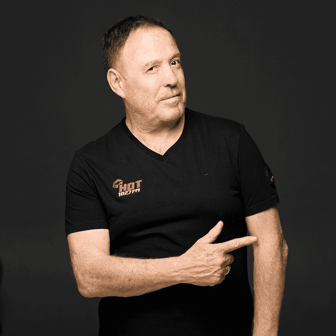 A man with short dark hair wearing a black t-shirt with a Hot 102.7 FM logo stands against a dark background, smiling slightly and pointing to his right with his index finger.
