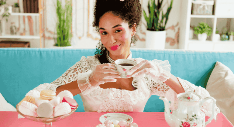 A woman in an elaborate white lace dress, gloves, and feathered hat sits at a pink table, smiling while holding a tea cup. There are pastries, a teapot, and cake in front of her, with a blue couch in the background.