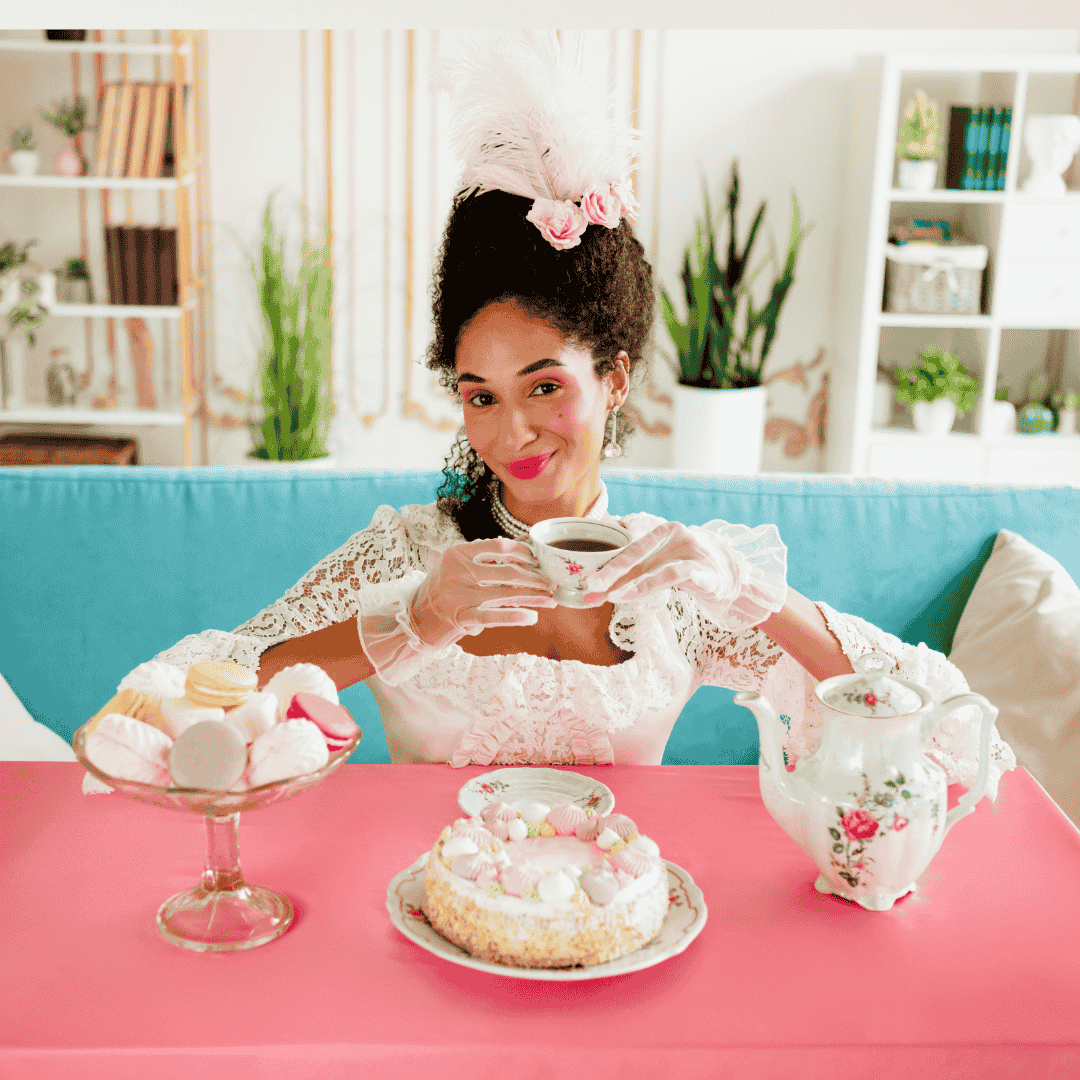 A woman in an elaborate white lace dress, gloves, and feathered hat sits at a pink table, smiling while holding a tea cup. There are pastries, a teapot, and cake in front of her, with a blue couch in the background.