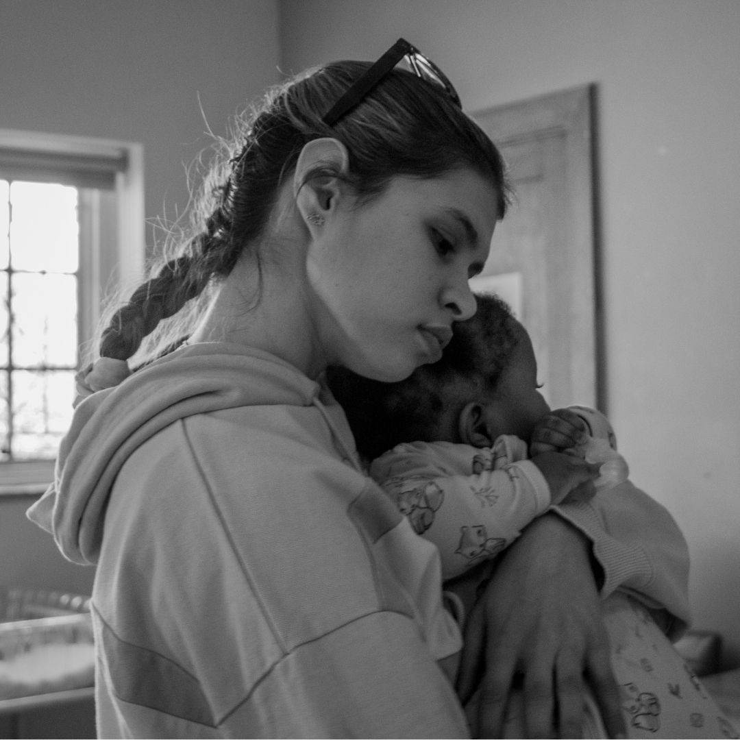 A young woman with a braid hugs and comforts a small baby dressed in patterned pajamas. Soft light enters through a window, giving the scene a calm, caring atmosphere—perfectly reflecting the spirit of Child Protection Week.