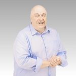 A smiling middle-aged man with short gray hair wears a light blue button-up shirt. He stands in front of a plain gray gradient background, clasping his hands together, ready to discuss the latest hot business trends.