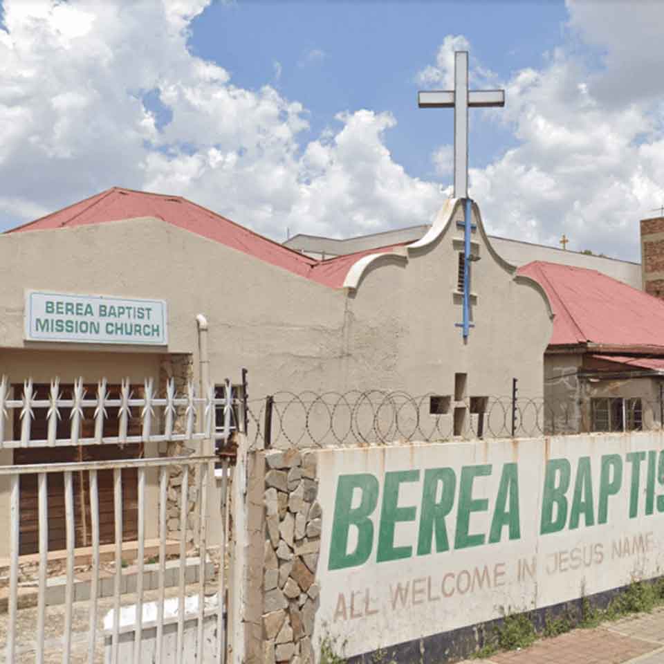 A beige building with a red roof, a white cross, and signs reading “Berea Baptist Mission Church” and “Berea Baptist—All welcome in Jesus name,” surrounded by a stone wall and metal fence under a cloudy sky.