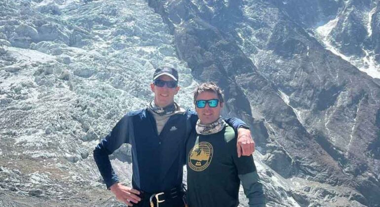 Two climbers wearing sunglasses and mountaineering gear stand arm-in-arm on a rocky, snowy mountainside with a towering, sunlit glacier and blue sky in the background.